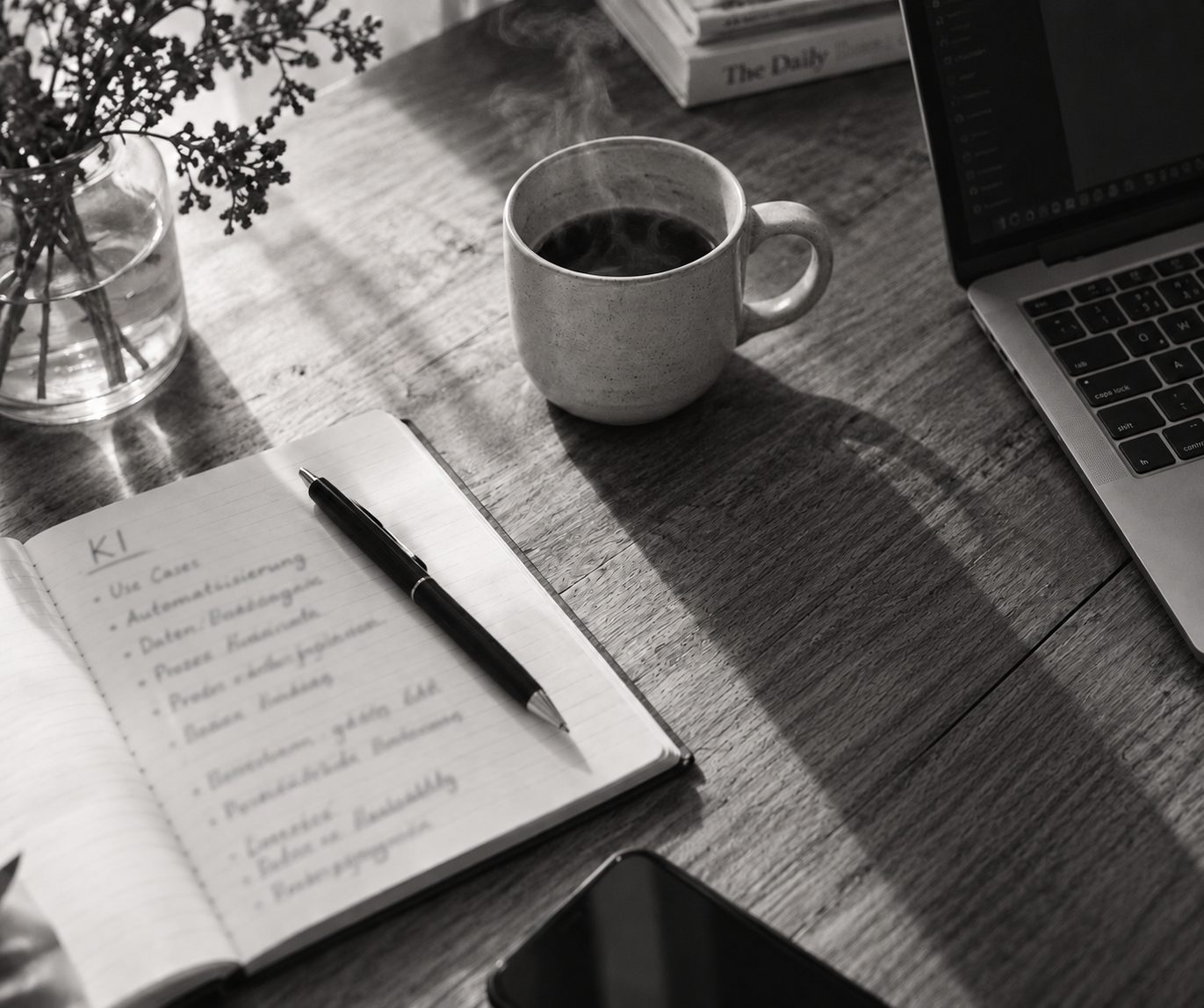 Holztisch mit Notizbuch, Stift, Kaffeetasse und Laptop im Morgenlicht.